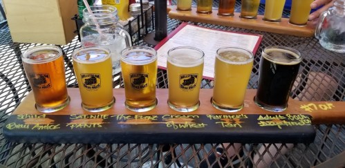 A wooden flight paddle with five different craft beers, labeled, on a table with a menu in the background.