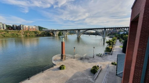 A scenic view of a river with a bridge, surrounded by greenery and buildings under a partly cloudy sky.