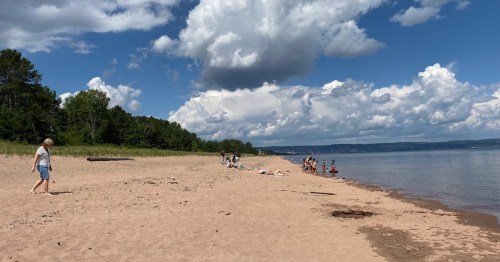 A sandy beach with people enjoying the sun, trees in the background, and a cloudy blue sky overhead.
