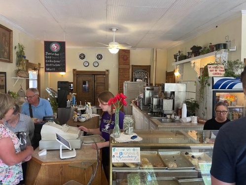 A cozy café interior with a counter, customers, and a barista serving drinks, surrounded by plants and baked goods.