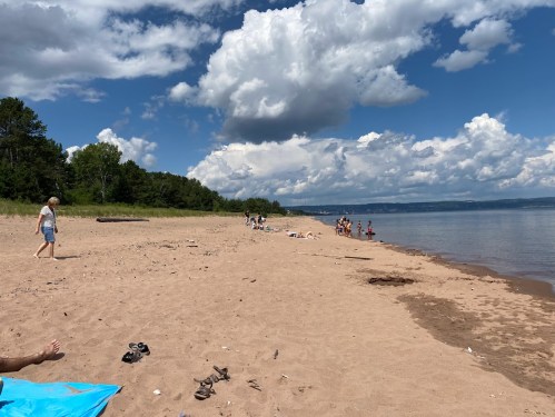 A sandy beach with people enjoying the sun, trees in the background, and fluffy clouds in a blue sky.