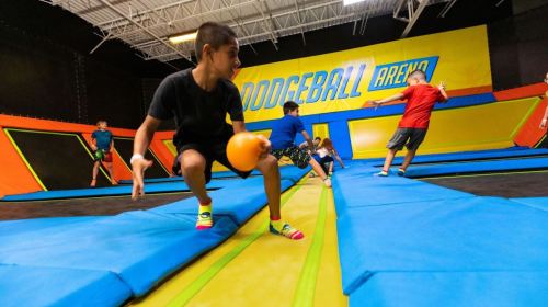 Kids playing dodgeball on trampolines in a colorful arena, with one child preparing to throw a ball.