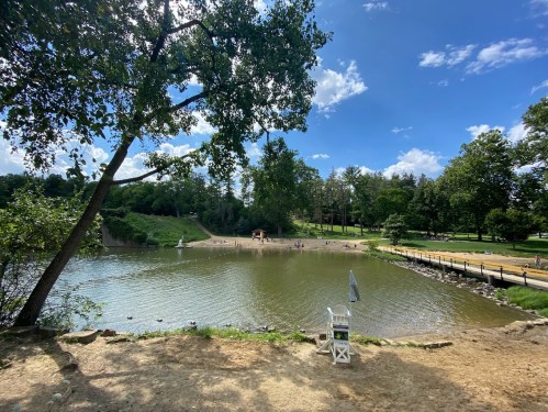 A serene park scene featuring a pond, sandy shore, trees, and people enjoying the outdoors under a blue sky.