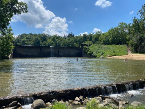 A serene lake with a waterfall, surrounded by lush greenery and people swimming on a sunny day.