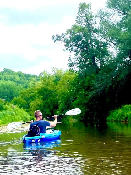 A person paddles a kayak on a calm river, with a dog sitting in the front, surrounded by lush greenery.