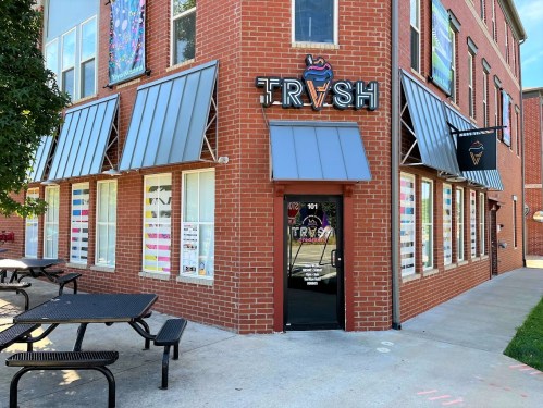 Exterior of a brick building with a sign reading "TRVSH," featuring large windows and outdoor seating.