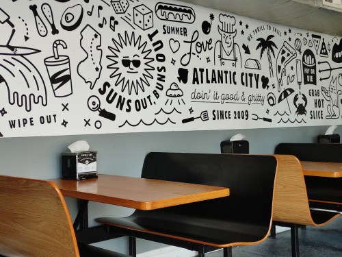A black and white mural featuring summer-themed illustrations on a restaurant wall, with empty wooden tables in front.