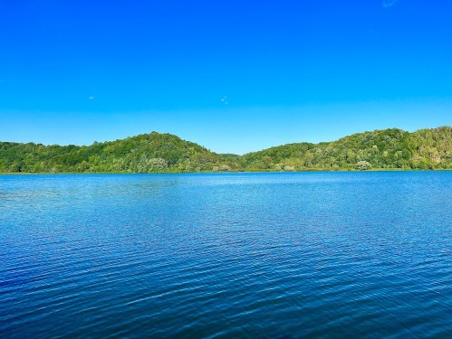 A serene lake surrounded by lush green hills under a clear blue sky. Gentle ripples on the water's surface.