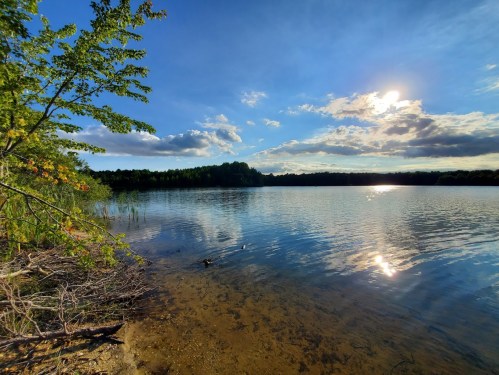 A serene lake scene with clear blue skies, fluffy clouds, and sunlight reflecting on the water's surface.