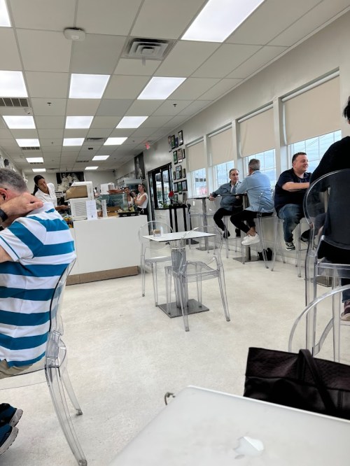 A bright café interior with clear chairs, tables, and customers chatting. A counter is visible in the background.
