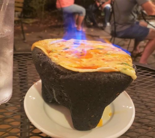 A sizzling dish on a black stone base, topped with melted cheese and flames, served on a white plate.