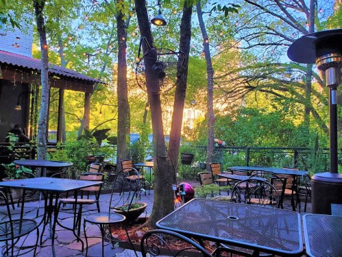A cozy outdoor patio with metal tables and chairs surrounded by trees and greenery, illuminated by soft evening light.