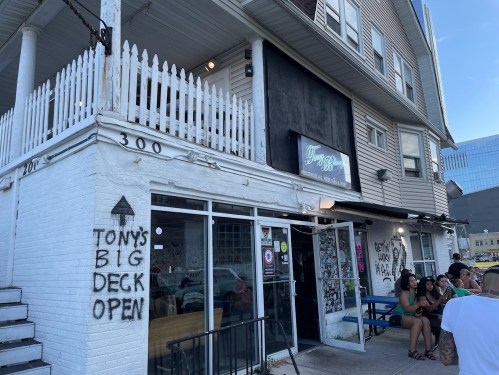 Exterior of a building with a sign for "Tony's Big Deck Open," featuring a white facade and people sitting outside.