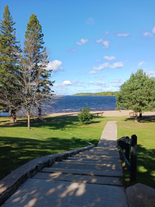 A pathway leads down to a serene lake, surrounded by trees and a clear blue sky with scattered clouds.