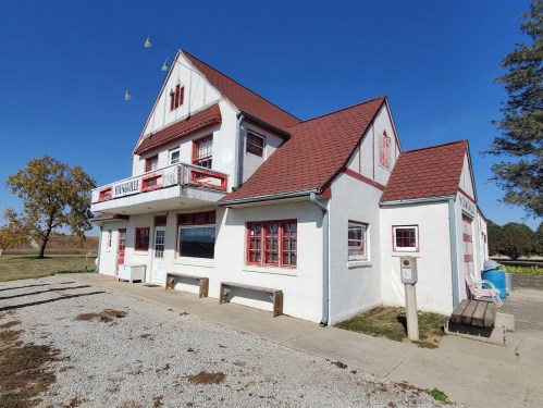 A white building with red accents and a sloped roof, featuring a balcony and surrounded by a gravel path and trees.