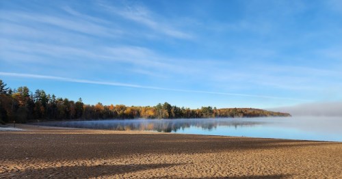 A serene lakeside view with a sandy beach, surrounded by trees and a misty horizon under a clear blue sky.