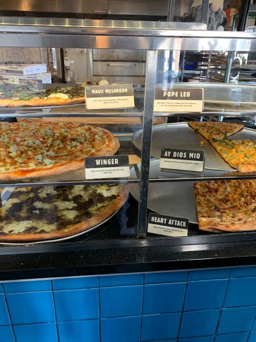 A display case with various pizza slices labeled: Magic Mushroom, Pope Leo, Winger, Ay Dios Mio, and Heart Attack.
