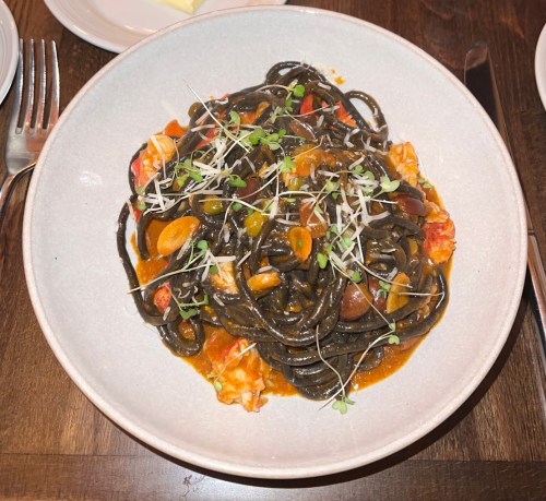 A bowl of black spaghetti with seafood, garnished with herbs and sliced garlic, served on a wooden table.
