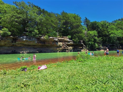 A sunny day at a green river with people swimming and playing near a rocky shore surrounded by trees.