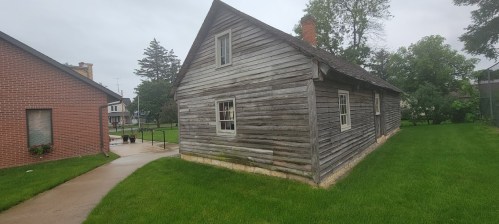 A weathered wooden house with a sloped roof, surrounded by green grass and trees, on a cloudy day.