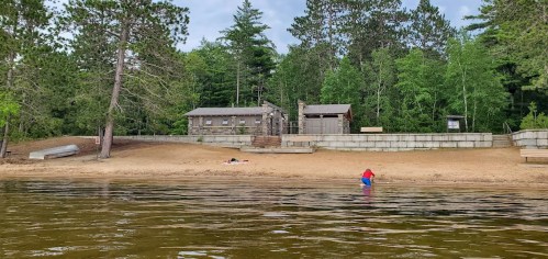 A sandy beach with a cabin and trees in the background; a person wades in the water near the shore.