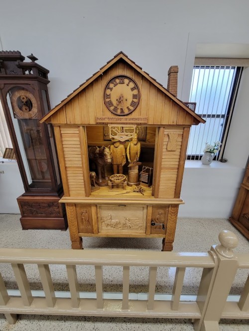 A wooden clock display featuring intricate carvings of two figures and various tools, set in a house-like structure.