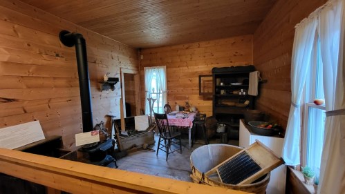 Cozy wooden interior of a rustic room with a stove, table, chairs, and vintage kitchen items. Natural light from windows.