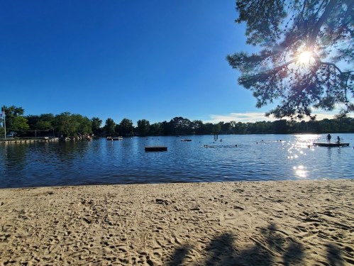 A serene lake scene with people paddling, surrounded by trees and a bright sun reflecting on the water.