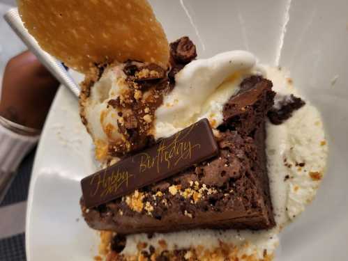 A slice of chocolate cake topped with ice cream, a cookie, and a "Happy Birthday" chocolate plaque.