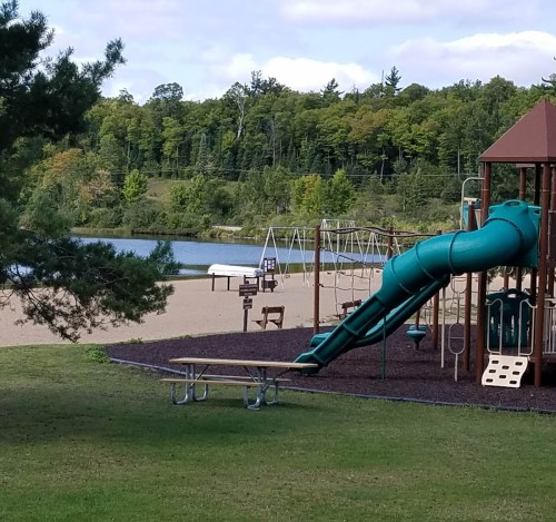 A playground with a slide and swings near a sandy area by a lake, surrounded by trees and a cloudy sky.