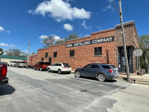 A brick building with "Iowa Distilling Company" sign, parked cars along the street, and a clear blue sky.