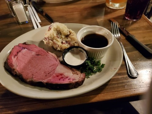 A plate with a slice of prime rib, mashed potatoes, and dipping sauces, garnished with parsley on a wooden table.