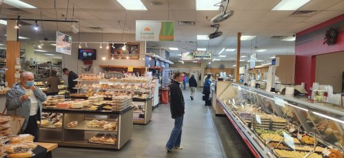 A bustling market interior with shelves of baked goods, salads, and customers browsing the aisles.