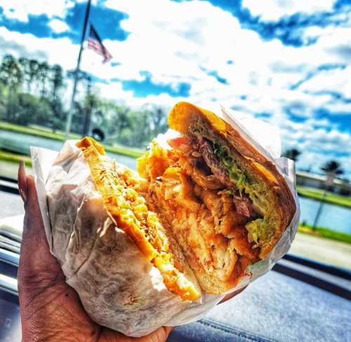 A hand holding a large, layered sandwich with chicken, lettuce, and tomato against a scenic background.