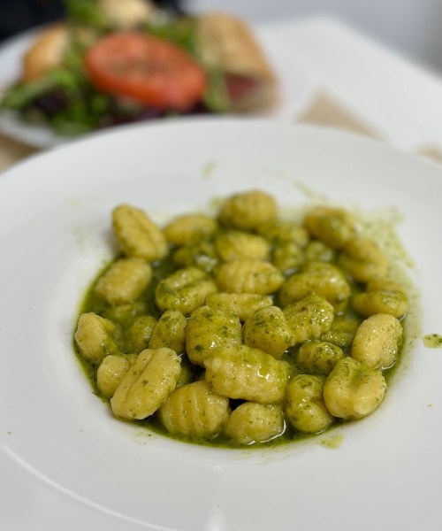A plate of gnocchi coated in green pesto sauce, with a side salad in the background.