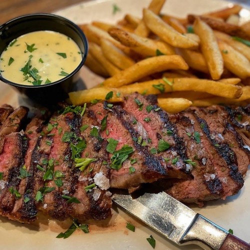 A plate featuring grilled steak garnished with parsley, served with French fries and a small cup of creamy sauce.