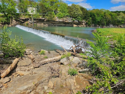 A serene river scene with a small waterfall, surrounded by lush greenery and rocky terrain under a clear blue sky.