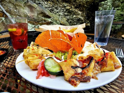A colorful plate of Mexican food featuring tacos, tamales, and grilled vegetables, with drinks on a table outdoors.