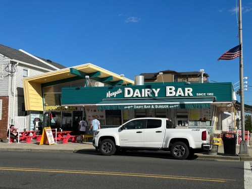 A retro-style dairy bar with a green awning, outdoor seating, and a white truck parked in front on a sunny day.