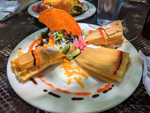 A plate with three tamales, a taco, black beans, guacamole, and colorful sauces on a black table.