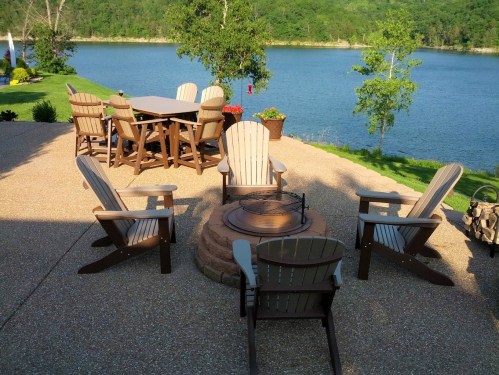 A lakeside patio with a dining table, chairs, and a fire pit surrounded by greenery and water.