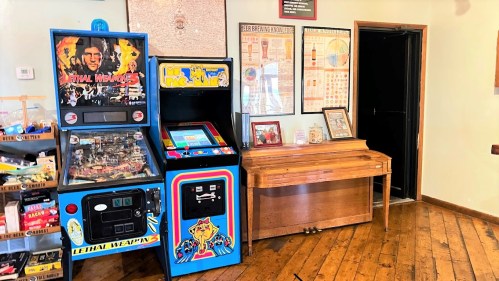 A cozy room featuring vintage arcade machines and a wooden piano against a wall with framed posters.