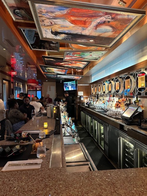 A stylish bar with colorful ceiling art, illuminated bottles, and patrons enjoying drinks in a vibrant atmosphere.