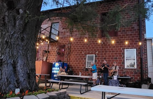 A band performs outdoors near a brick building, with string lights and picnic tables in a lively atmosphere.