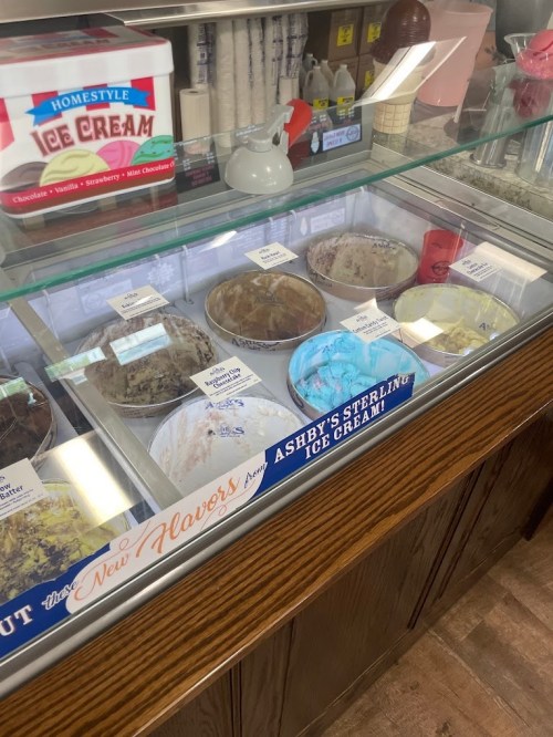 A display case filled with various ice cream flavors, labeled with names, at Ashby's Sterling Ice Cream shop.