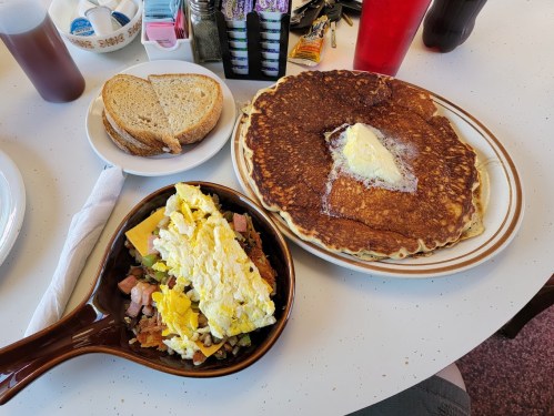 A plate with a large pancake topped with butter, a bowl of scrambled eggs with ham, and two slices of bread on the side.