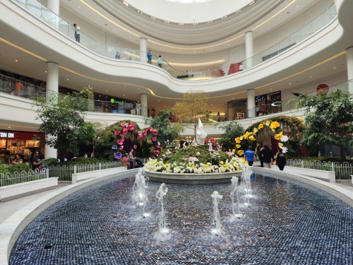 A spacious mall interior featuring a fountain, colorful flowers, and shoppers on multiple levels.