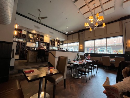 Interior of a restaurant featuring tables, a bar area, and large windows with a view of the street outside.