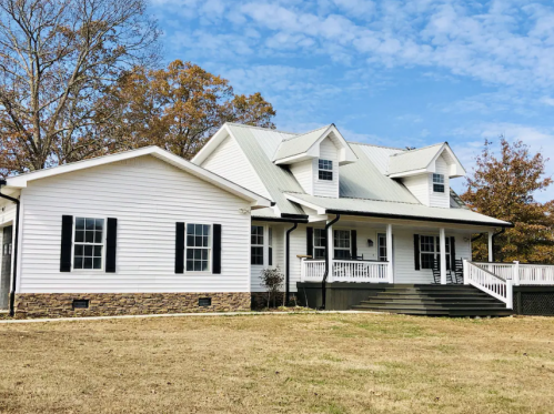 A charming white house with a metal roof, black shutters, and a spacious front porch, set against a blue sky.