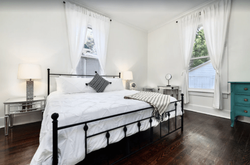 Bright bedroom with a black metal bed, white linens, and large windows, featuring a turquoise dresser and modern decor.